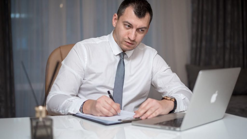 Focused businessman in white shirt writing notes at desk with laptop.