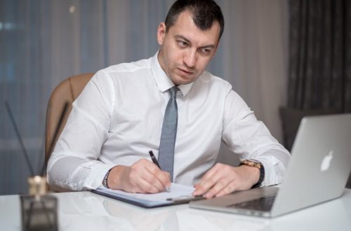 Focused businessman in white shirt writing notes at desk with laptop.
