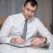 Focused businessman in white shirt writing notes at desk with laptop.